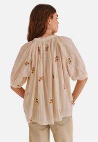 Lys beige bluse med puffede ærmer og blomsterbroderi. Stoffet er gennemsigtigt med en struktureret overflade og har samlet detalje bagpå.