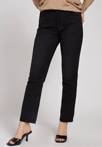 Jeans in denim nero con una vestibilità a gamba dritta, dotati di tasche anteriori e una texture liscia. Abbinati a tacchi aperti neri.
