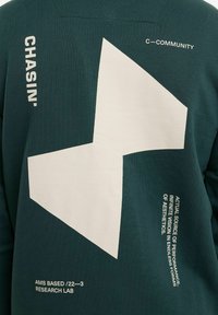 Felpa verde scuro con un grande design geometrico bianco e testo. Il tessuto ha una finitura testurizzata dallo stile casual.