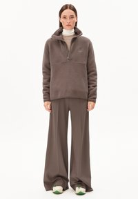 Bruine fleece pullover met een hoge kraag en rits, gecombineerd met brede bruine broeken. Model draagt schoeisel met groene accenten.