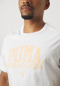 Bela bombažna majica z velikim, zbledelim oranžnim potiskom "PUMA sports club" in majhnim logotipom, s klasičnim krojem ovratnika.