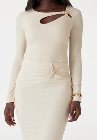 Femme portant une robe moulante beige à manches longues, avec des découpes ornées de boutons dorés au niveau de la poitrine et de la taille, et un bracelet doré.