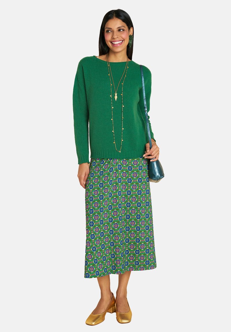 Frau mit grünem Pullover, grünem Blumenkleid, goldenen Heels und langen Halsketten, die eine metallisch türkisfarbene Schultertasche hält, lächelt und nach rechts schaut.