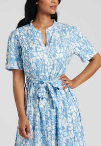 Niebieska midi sukienka w kwiaty z białymi wzorami, krótkimi rękawami, zapięciem na guziki oraz akcentem w postaci związanej talii. Miękki materiał z płynącą sylwetką.