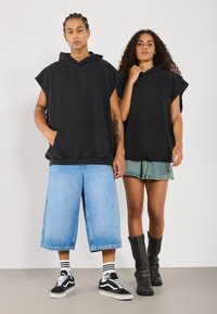 Čierne oversized kapucňové tielka bez rukávov, modré voľné denims šortky a zelená denimová sukňa. Obuv zahŕňa čierne čižmy a tenisky Vans.