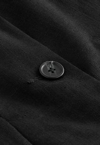 Gros plan d'un bouton noir à quatre trous cousu sur un tissu noir texturé avec des coutures visibles et de légers plis dans le tissu.