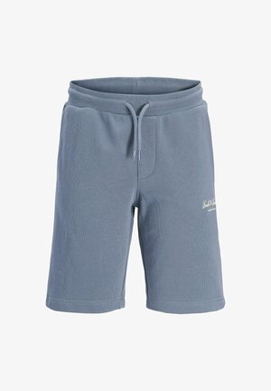Chico adolescente de pie, vestido con camiseta negra, pantalones cortos azul claro, calcetines blancos con texto y zapatillas blancas con franjas negras, sobre un fondo liso.
