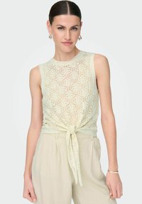 Top de punto sin mangas en verde claro, con un patrón de encaje y un detalle de lazo anudado en la cintura, combinado con pantalones de talle alto beige.