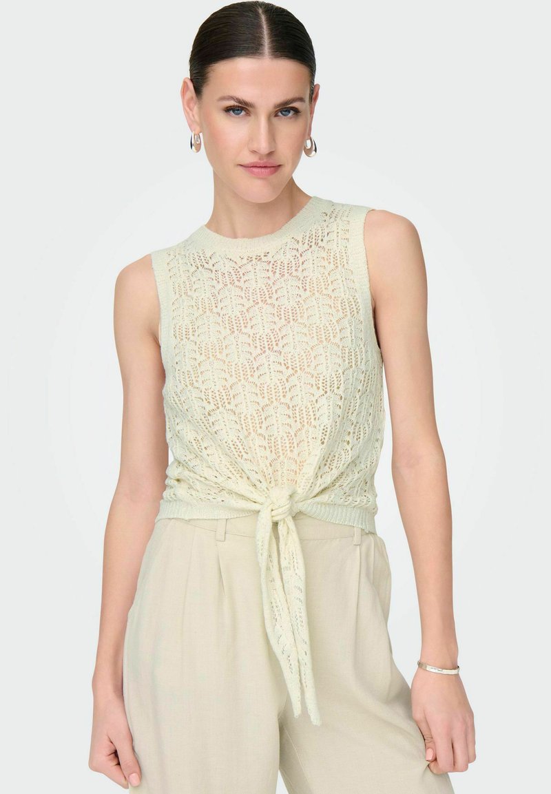 Top de punto sin mangas en verde claro, con un patrón de encaje y un detalle de lazo anudado en la cintura, combinado con pantalones de talle alto beige.