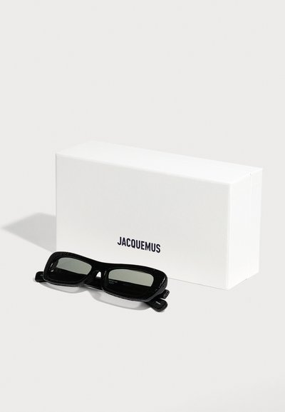 Lunettes de soleil rectangulaires noires avec monture texturée placées devant une boîte blanche de la marque Jacquemus sur un fond uni.