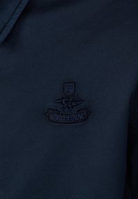 Tissu bleu foncé avec un emblème cousu représentant un oiseau, un bouclier et le texte « FAÇONNABLE » sous le motif.