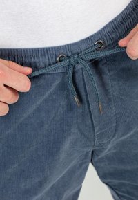 Pantalones de pana azul con cierre de cordón. La cinturilla cuenta con ojales de metal y la tela tiene una textura acanalada.
