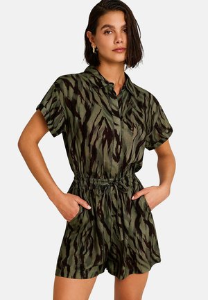 Femme portant une combinaison courte à manches courtes, imprimé animal vert et noir, avec col et taille à cordon, les mains dans les poches, face à l'avant.