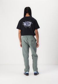 Tricou negru cu un design grafic pe spate, asortat cu pantaloni din catifea verde și adidași albastre cu dungi.