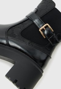 Bottine en cuir noir avec motifs embossés, panneau latéral élastique, sangle à boucle dorée et talon bloc épais sur fond blanc.