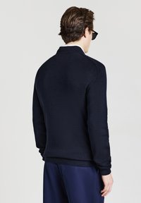Maglione navy a maglia con colletto piatto e polsini a coste. Texture liscia con silhouette aderente, indossato con pantaloni navy abbinati.