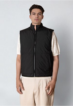 Uomo in piedi che indossa un gilet nero senza maniche con cerniera sopra una camicia beige chiaro a maniche corte e pantaloni beige su uno sfondo semplice.