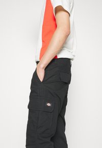 Personne portant une chemise blanche et orange à manches courtes et un pantalon cargo noir, les mains dans les poches, sur un fond blanc uni.