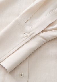 Light beige shirt with a smooth texture, featuring a single button at the cuff and a rolled sleeve design. Simple stitching detail visible.