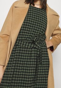 Abrigo color camello combinado con un vestido de cuadros en verde oscuro y negro, que cuenta con una cinturilla atada y una tela texturizada.