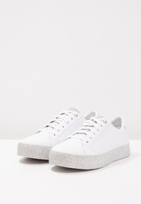 Sneakers bianche con tomaia in pelle liscia, lacci piatti e una suola a piattaforma argentata scintillante. Branding minimale vicino al tallone.