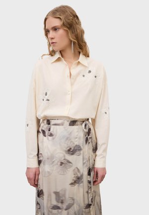 Vaaleahiuksinen nainen, jolla on laineikkaat vaaleat hiukset, pukeutuneena kermaiseen paitaan, jossa on kukka-ompeluita, ja vaaleanharmaaseen kuvioituun kietaisuhameeseen, seisoo yksinkertaisen taustan edessä.