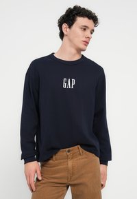 Μπλε ναυτικό πουκάμισο με μακριά μανίκια και λευκό λογότυπο "GAP" στο στήθος, άνετη εφαρμογή, ριμπ λαιμόκοψη, σε συνδυασμό με ανοιχτό καφέ παντελόνι από κορδούρα.