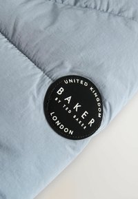 Tissu de doudoune bleu clair avec une étiquette circulaire noire cousue portant le mot "BAKER" de Ted Baker, avec une inscription blanche et une texture lisse.
