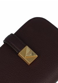 Portefeuille en cuir bordeaux avec une finition texturée, doté d'un fermoir triangulaire en ton doré avec le nom de la marque gravé et d'une sangle marron foncé.