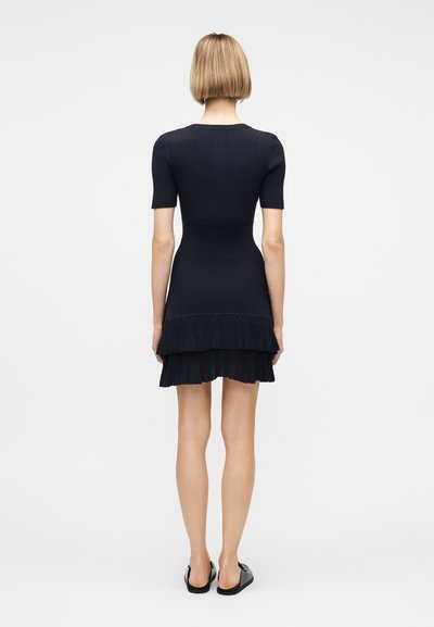 Γυναίκα με κοντά ξανθά μαλλιά, ντυμένη με μαύρο πλεκτό φόρεμα με κοντά μανίκια και φούστα με στρώσεις βολάν, που στέκεται με την πλάτη γυρισμένη σε λευκό φόντο.