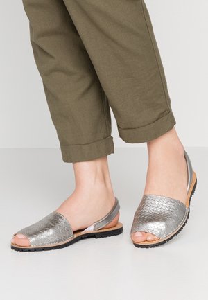 Stopy w metalicznie srebrnych, plecionych sandałach typu slingback w połączeniu z podwiniętymi oliwkowozielonymi spodniami na jednolitym tle.