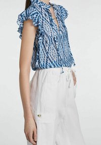 Blouse semi-transparente à motif bleu et blanc, avec manches à volants et col avec collerette. Associée à un pantalon blanc ample avec taille à cordon.
