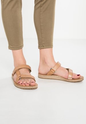 Voeten met beige open-toe sandalen met twee bandjes en rode nagellak, gecombineerd met kortere lichtbruine broeken.