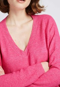 Pull à col en V rose avec une texture douce, des manches longues et des poignets côtelés. Le matériau semble léger et légèrement duveteux.