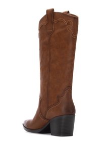 Botas de vaquero de cuero marrón con costuras decorativas, una punta ligeramente puntiaguda, un caña alta y un tacón apilado. Con un borde superior ondulado.