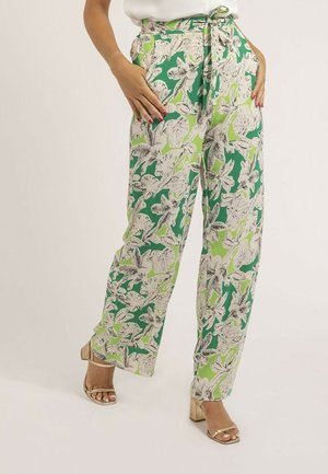 Femme portant un pantalon à jambes larges à fleurs vertes et blanches avec des poches latérales et des sandales à talons blocs dorées à bout ouvert, les mains reposant dans les poches.