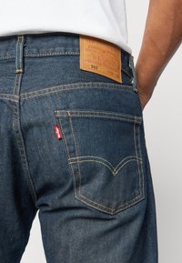 Jeans de mezclilla azul oscuro con un parche de cuero en la cintura trasera, con costuras naranjas y una etiqueta roja en el bolsillo trasero.