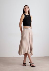 Top negro ajustado sin mangas, falda maxi beige con pliegues y una cinturilla metálica. Modelo lleva sandalias de tacón negro y está de pie sobre un fondo neutro.