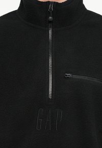 Μαύρο φλις πουλόβερ με ημι-φερμουάρ και μικρή οριζόντια τσέπη με φερμουάρ στο στήθος, με ανάγλυφο λογότυπο GAP κάτω από το φερμουάρ.