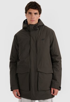 Man wearing dark green hooded winter jacket with two large front pockets and logo on chest, neutral background.