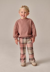 Vaaleanpunainen huppari puffisolmuilla, yhdistettynä ruudullisiin leveälahkeisiin housuihin, joissa on vaaleanpunaista, beigeä ja mustaa. Vaaleanruskeat slip-on-kengät täydentävät asua.