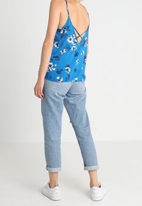 Persona che indossa una canotta floreale blu con dettagli incrociati sulla schiena, jeans azzurri a risvolto e sneakers bianche Reebok, in piedi con la schiena rivolta.