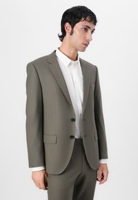 Olive green tailored suit with a single-breasted jacket, notch lapels, two front pockets, and black buttons, paired with a white shirt.