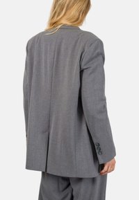 Blazer grigio su misura realizzato in tessuto testurizzato. Presenta una fessura centrale, polsini con quattro bottoni e una vestibilità rilassata, mostrato da dietro.