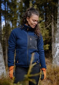 Navy fleece jacket with a black smooth fabric chest panel, zip pockets, and orange gloves. Textured and warm for outdoor use.