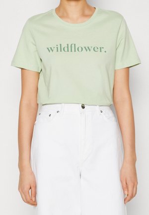 Osoba nosząca jasnozielony t-shirt z napisem „wildflower.” i białe spodnie z wysokim stanem na jednolitym tle.