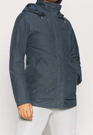 Personne portant une veste à capuche gris foncé avec les mains dans les poches et un pantalon blanc, debout devant un fond clair uni.