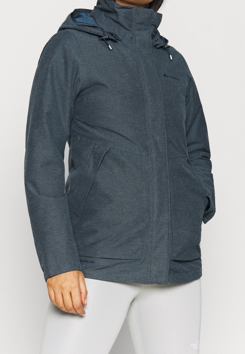 Personne portant une veste à capuche gris foncé avec les mains dans les poches et un pantalon blanc, debout devant un fond clair uni.