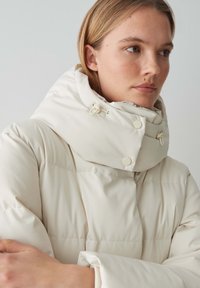 Veste en duvet crème avec un col haut, dotée de boutons-pression et de cordons réglables, présentant une texture lisse et brillante.