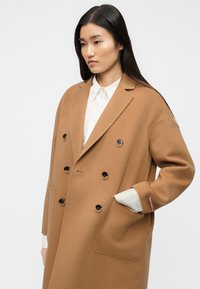 Παλτό διπλής σειράς σε καμηλό χρώμα από απαλό μείγμα μάλλινης ύφανσης, με μεγάλα μαύρα κουμπιά, πλαϊνές τσέπες και άνετη εφαρμογή. Φορεμένο πάνω από ένα λευκό πουκάμισο.
